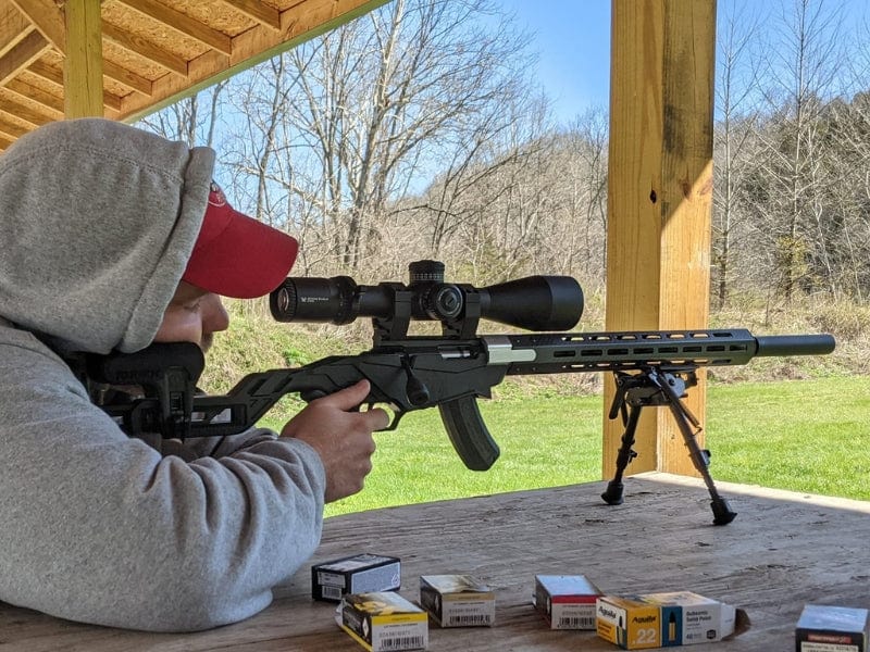 Person aiming a scoped rifle at an outdoor shooting range with ammunition boxes nearby