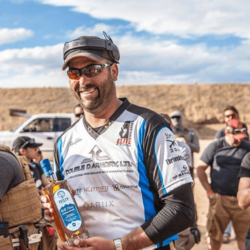 Smiling man at outdoor shooting event holding a bottle of liquor, wearing sunglasses and a sports jersey